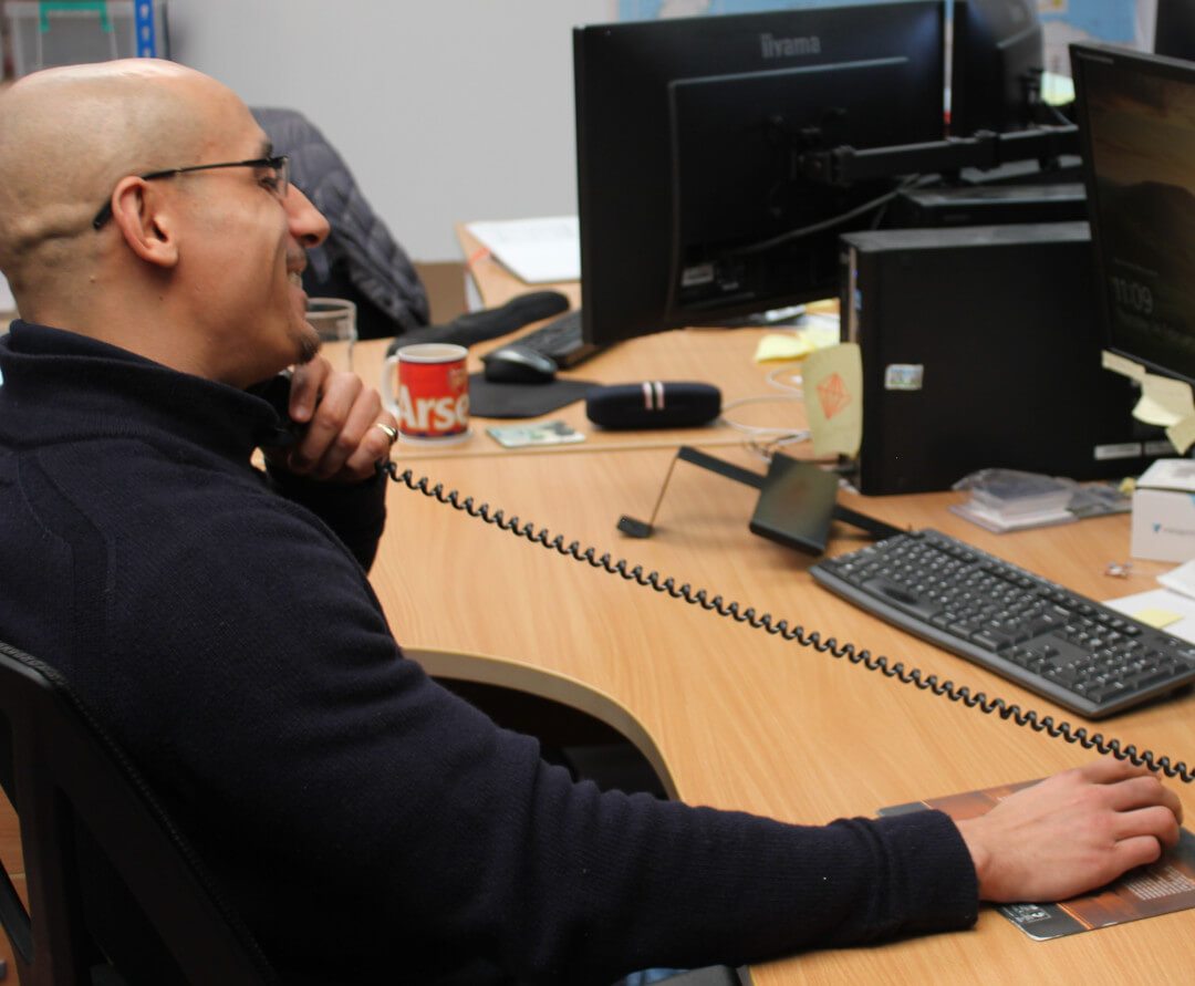 Profile of a smiling man sat at a desk with the phone to his ear while clicking his computer mouse to help illustrate GB Electronics' after sales service.